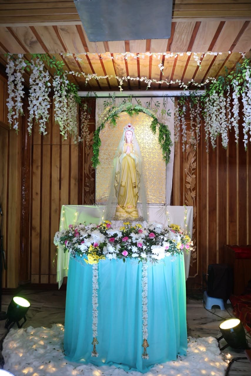 The statue of Our Lady standing at her shrine in the parish, under an arch of green leaves, with flowers banked on the altar below her